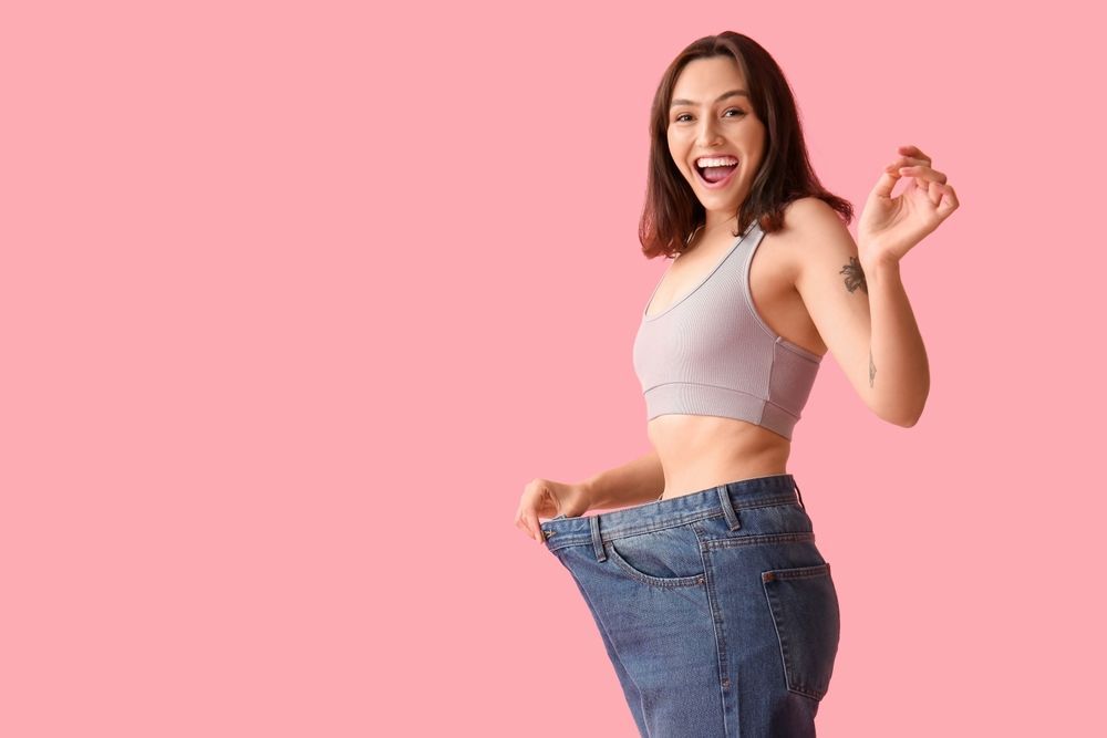A woman is wearing very large jeans on a pink background.