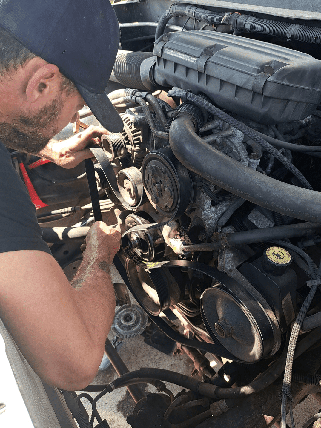 Man working on a car engine, inspecting belts and pulleys. Engine bay is open and illuminated by sunlight.