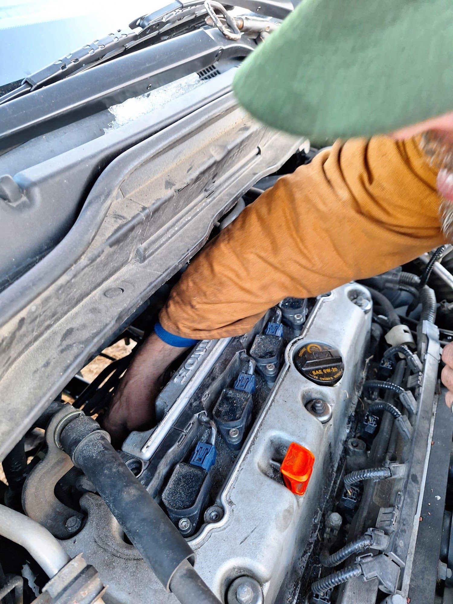 Person working on a car engine, reaching into the engine bay with an orange-sleeved arm.