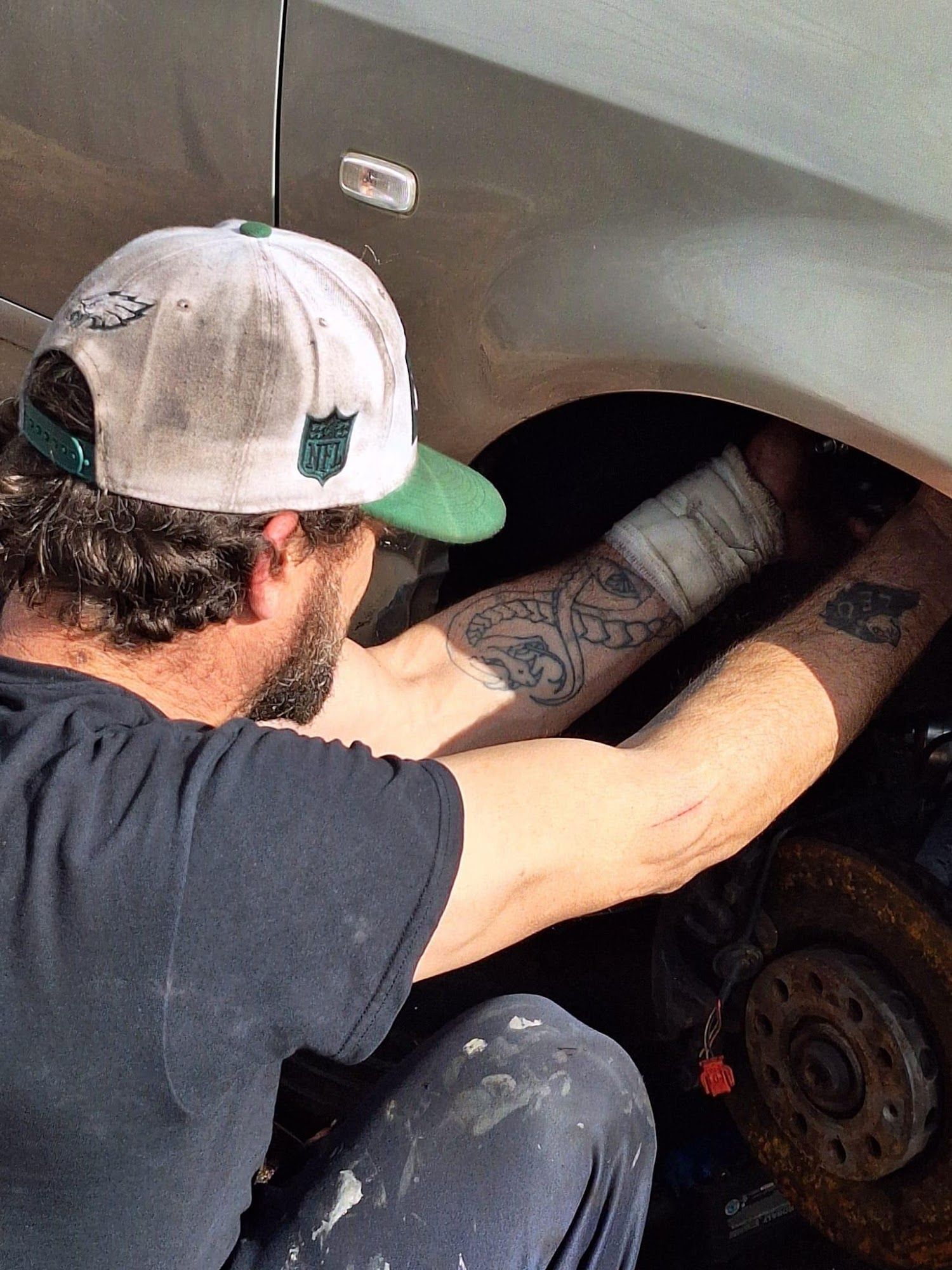 Man working on a car wheel, reaching into the wheel well. He wears a hat, and has a bandaged arm.