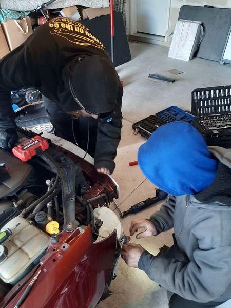 Two people in hoodies working on a car in a garage. Tools and car parts are visible.