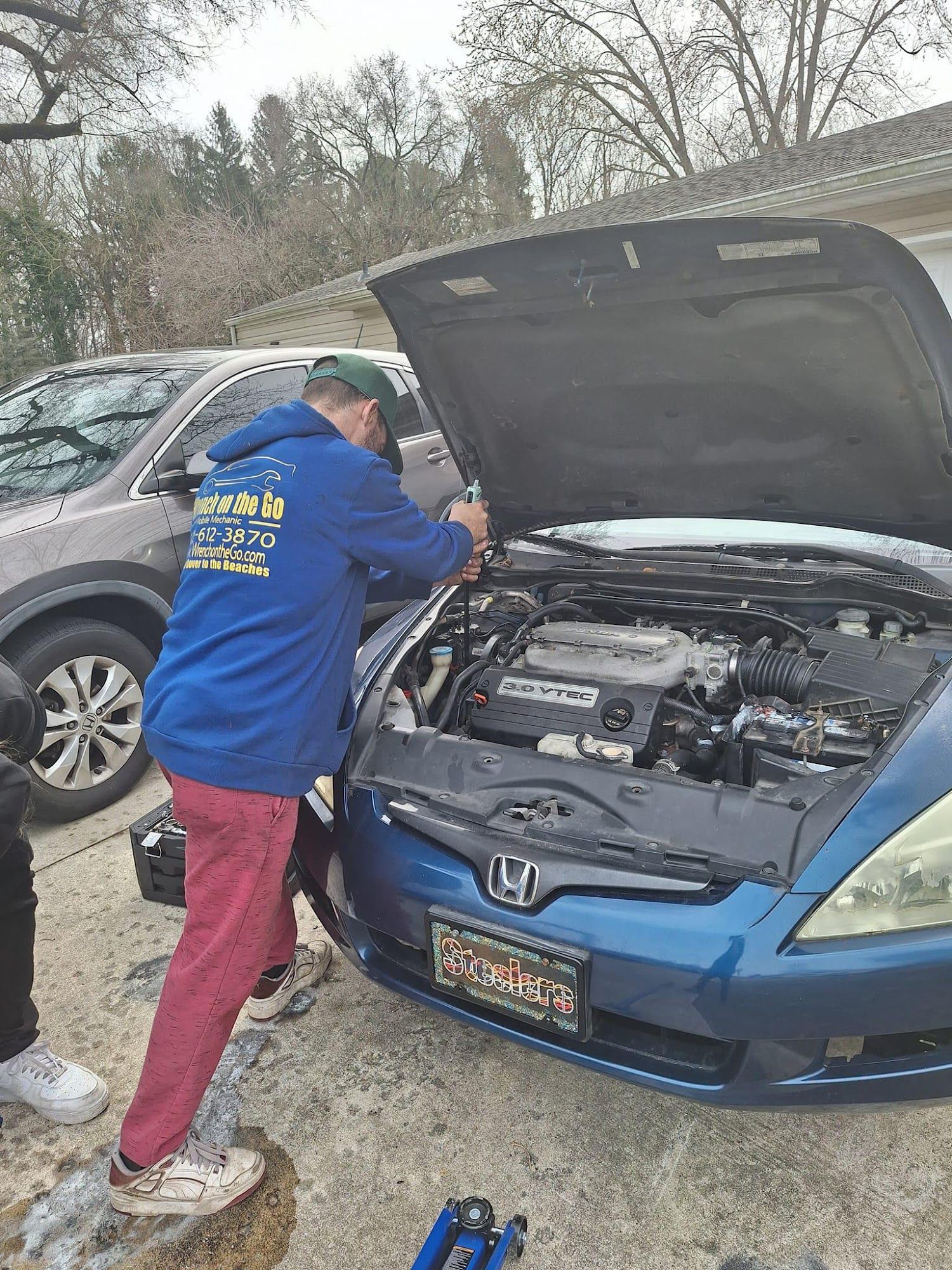 Person in blue jacket working on a blue car with the hood open in a driveway.
