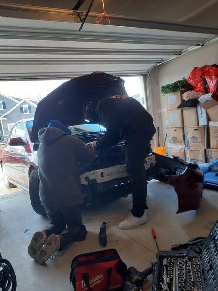Two people work on a car in a garage. One kneels, the other leans into the open hood. A detached bumper sits nearby.