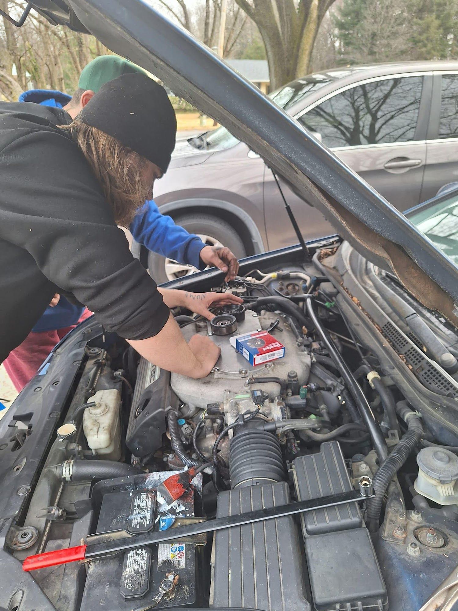 Two people working on a car engine under the open hood. One is wearing a black beanie.