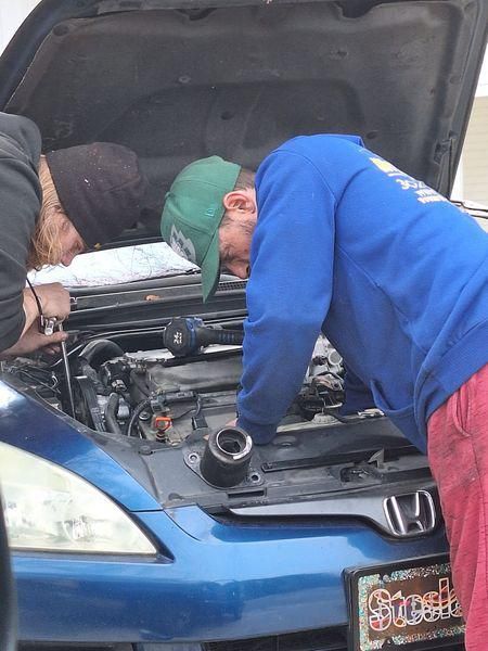 Two people working on a blue car engine. One wears a green hat and blue sweatshirt, the other a black hat.