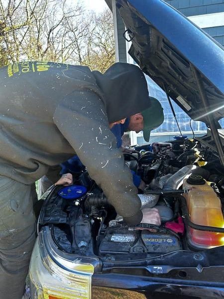 Person working on a car engine with hood open, wearing a hooded sweatshirt.