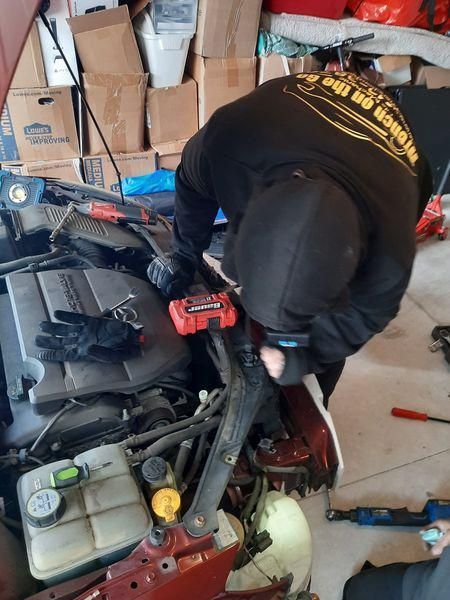 Person in a black hoodie working on a car engine in a cluttered garage, using tools.
