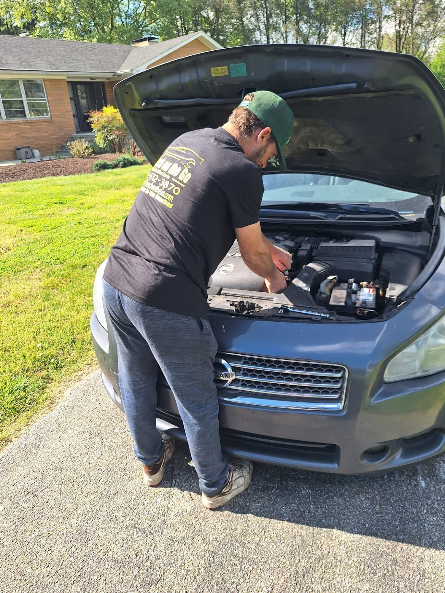 Man working on a car engine in a driveway, wearing a green cap and black shirt.