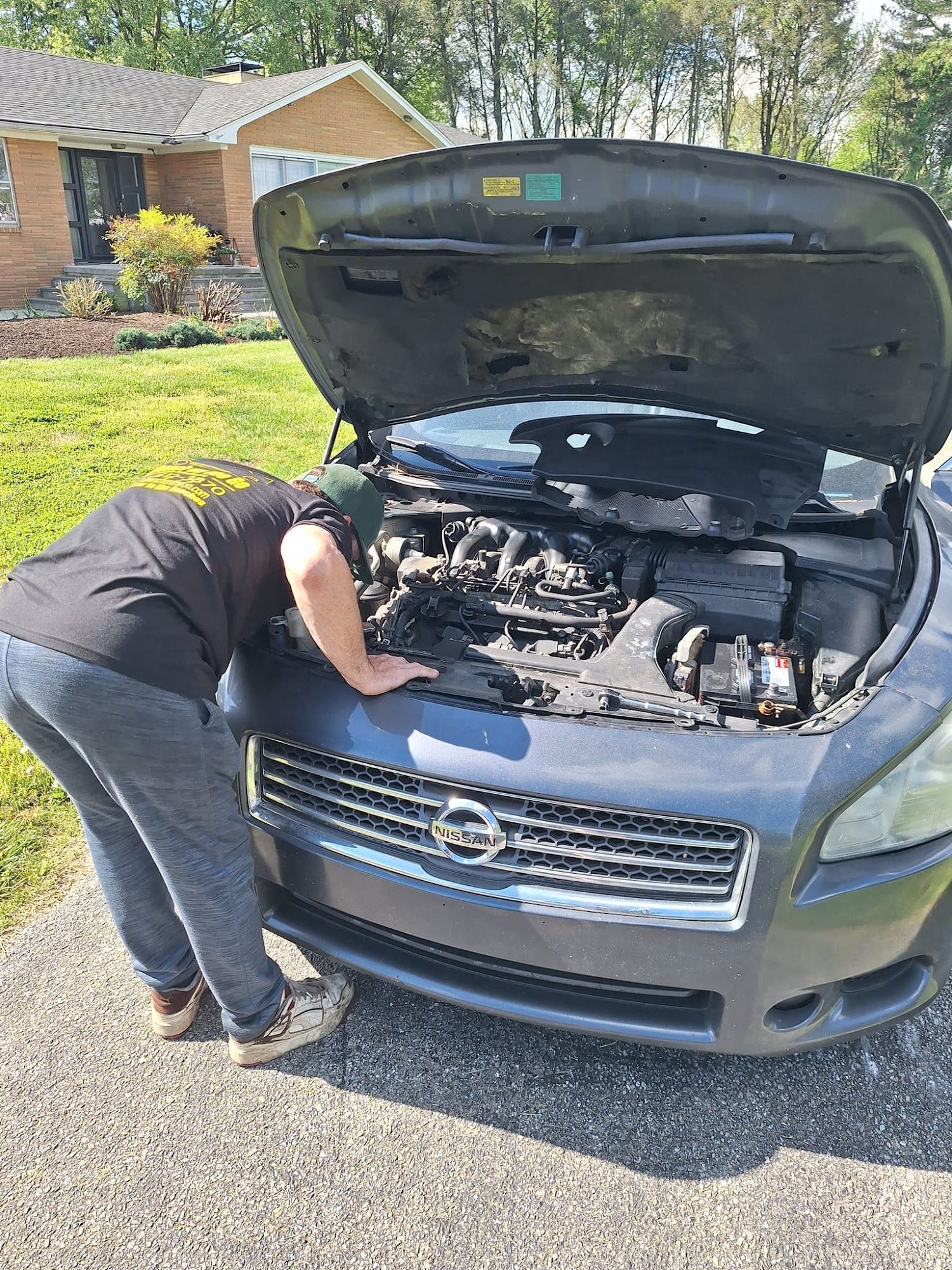 Person leans over a car with the hood open, examining the engine. Outdoors, sunny day.