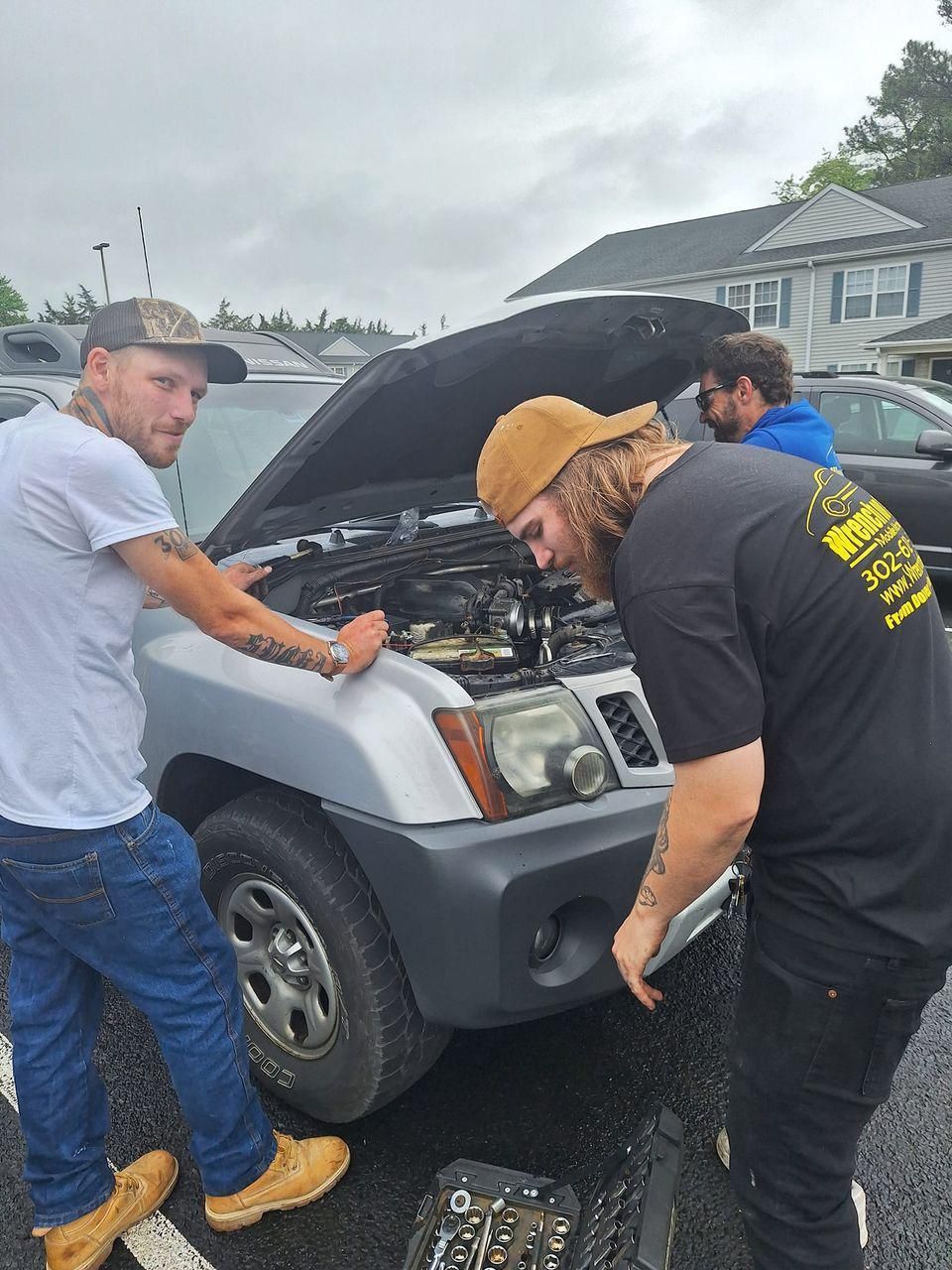 Three men working on a car with the hood open, parked in a lot under a cloudy sky.