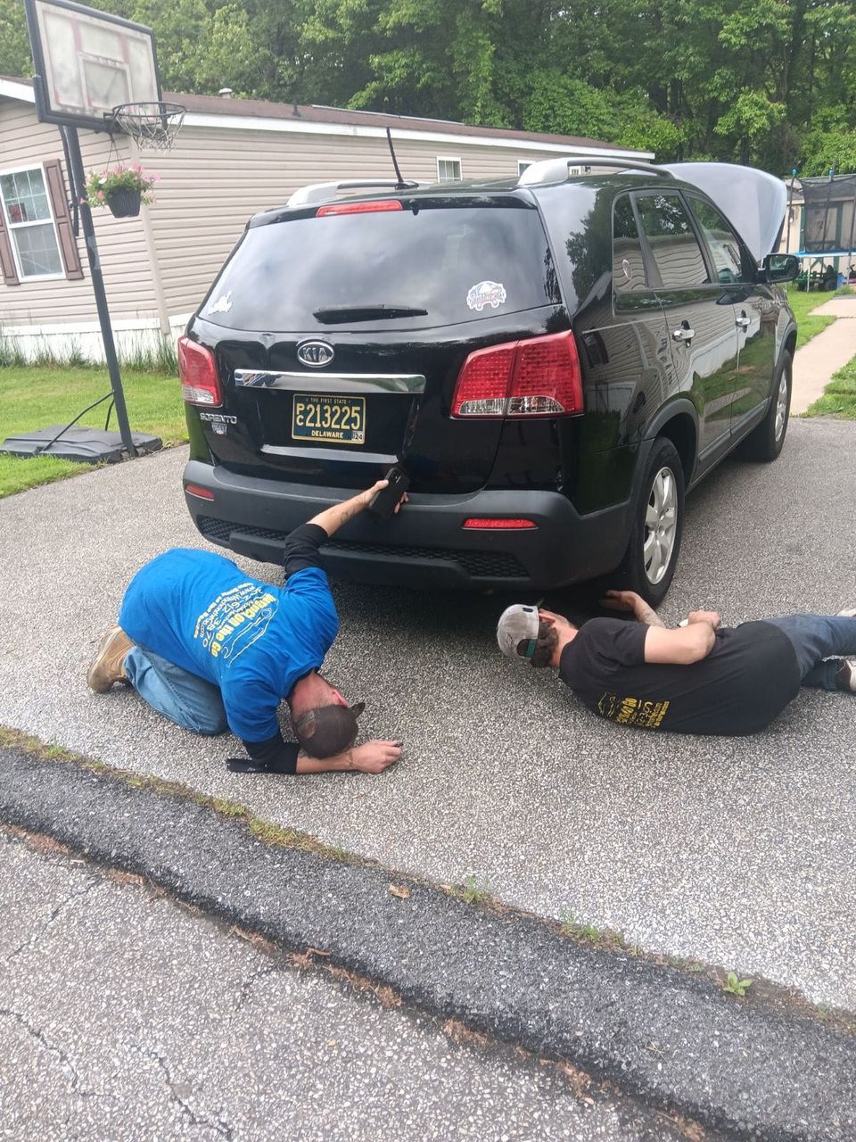 Two people working on a black SUV with the hood up, kneeling and lying on the ground in a driveway.