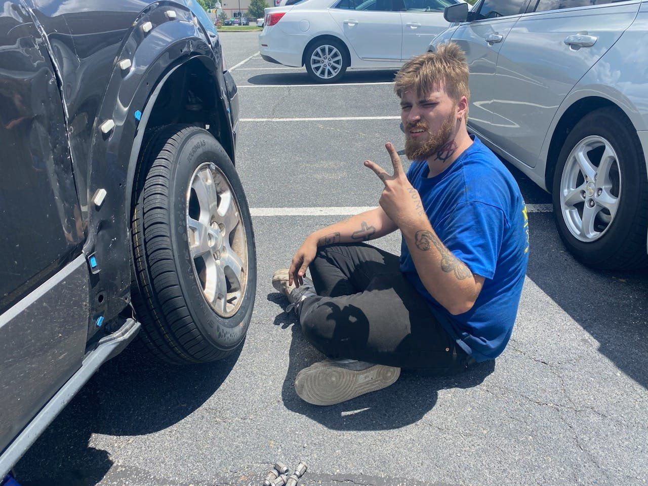Man sitting next to car with damaged fender. He's making a peace sign in a parking lot.