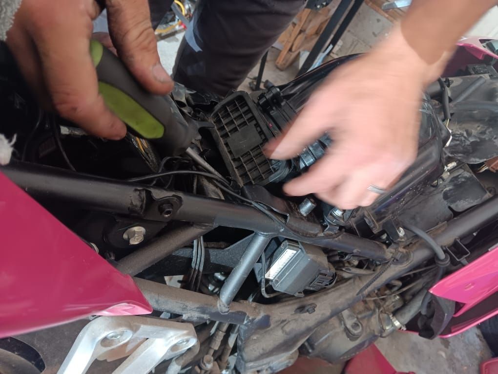 Hands working on a motorcycle, holding a tool and manipulating a black component, pink bodywork visible.