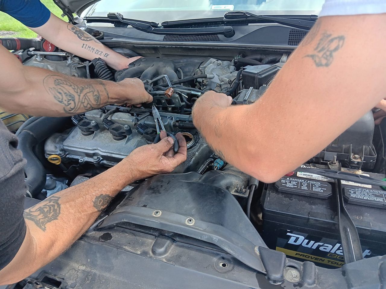 Three people working on a car engine; hands and arms are visible.