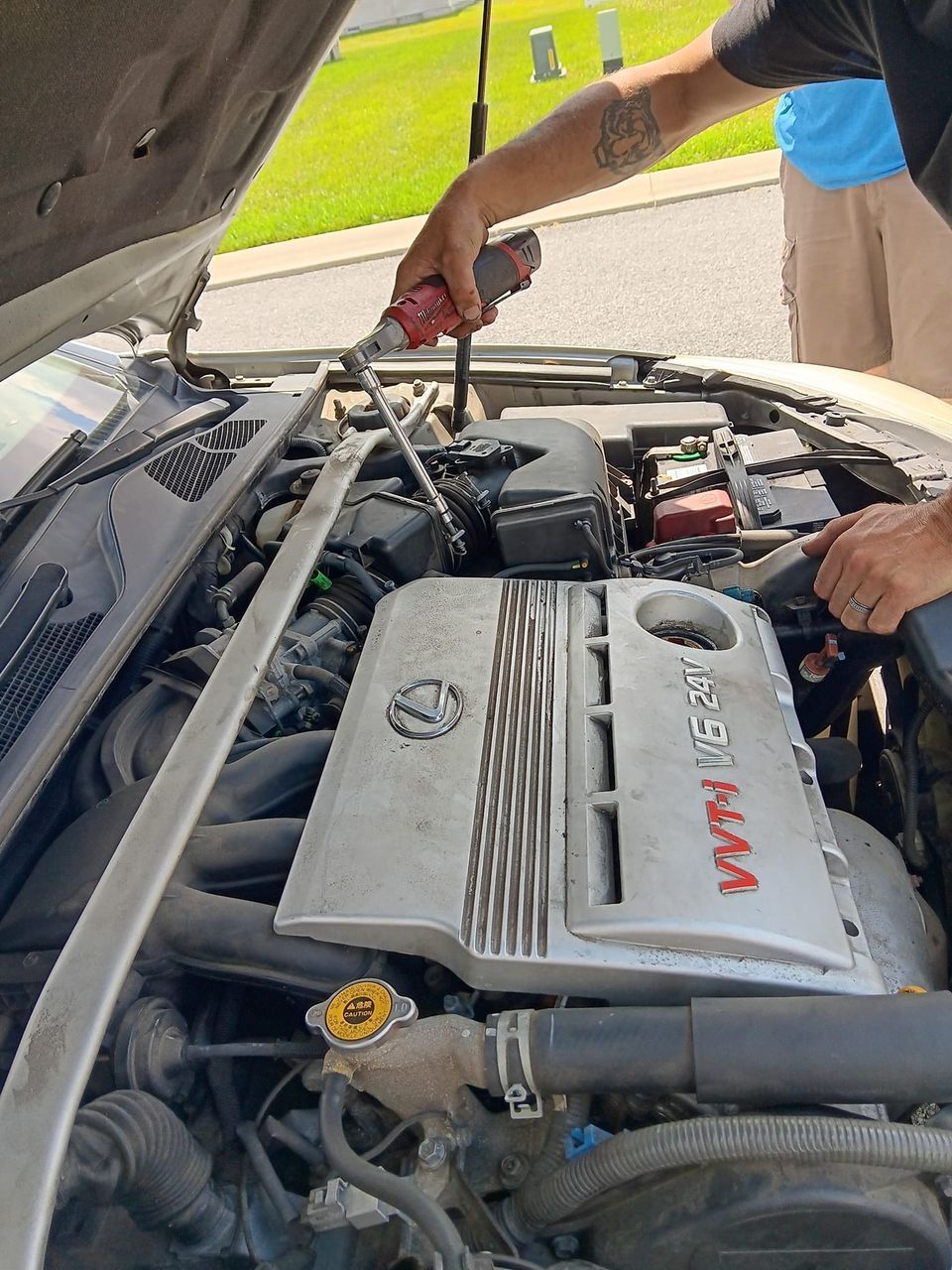Man working on a car engine, using a drill, outdoors. Car hood open, engine visible, gray color.