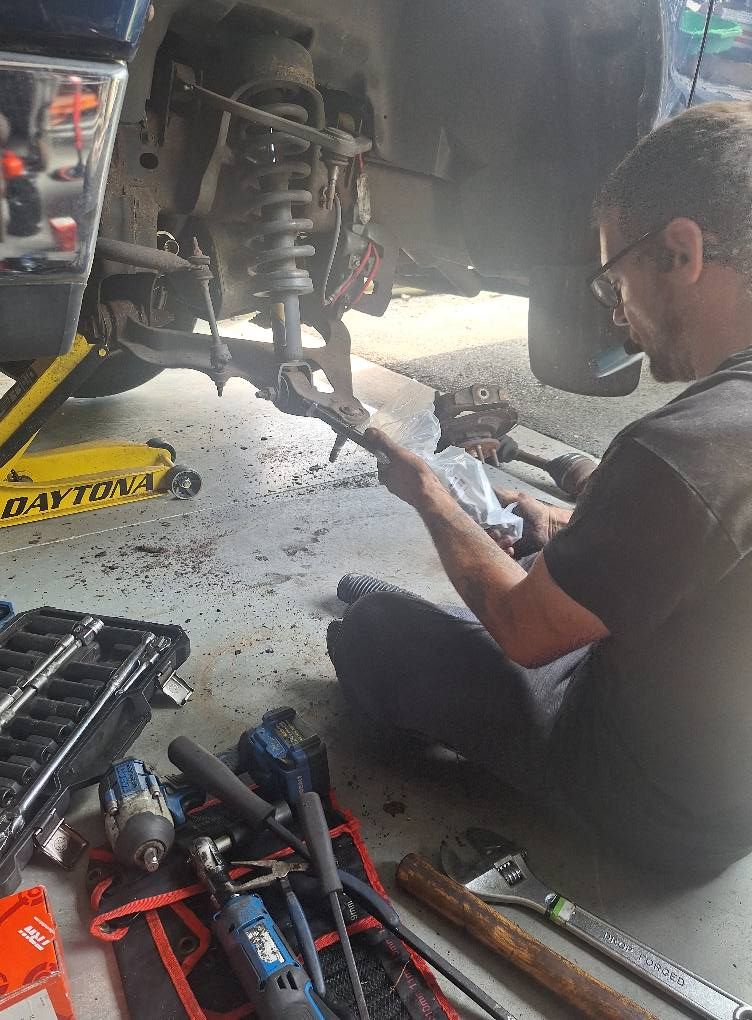 Man working on vehicle suspension in a garage, holding tool. Tools scattered on floor; jack supports car.