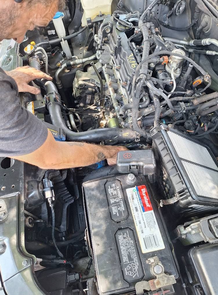 Person working on a car engine, hands visible. Car engine bay is open, battery is visible.