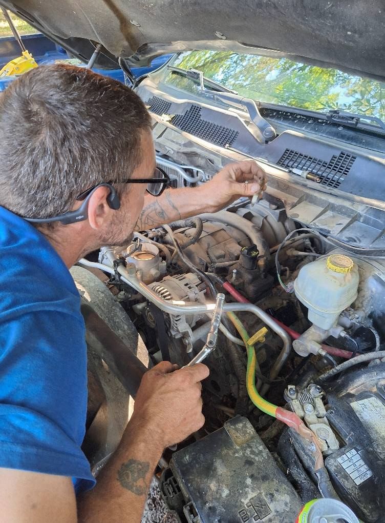 Man working on a car engine, holding pliers. Engine bay open, outdoors, sunny.
