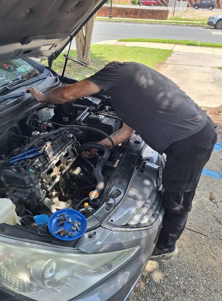 Person working on a car engine outdoors. Gray car, open hood. Tools and parts visible.