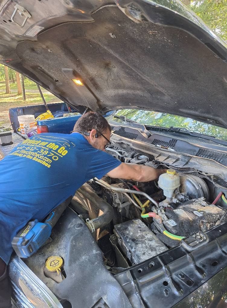 Mechanic works on a truck engine, outdoors. The hood is open; he wears a blue shirt with a company logo.