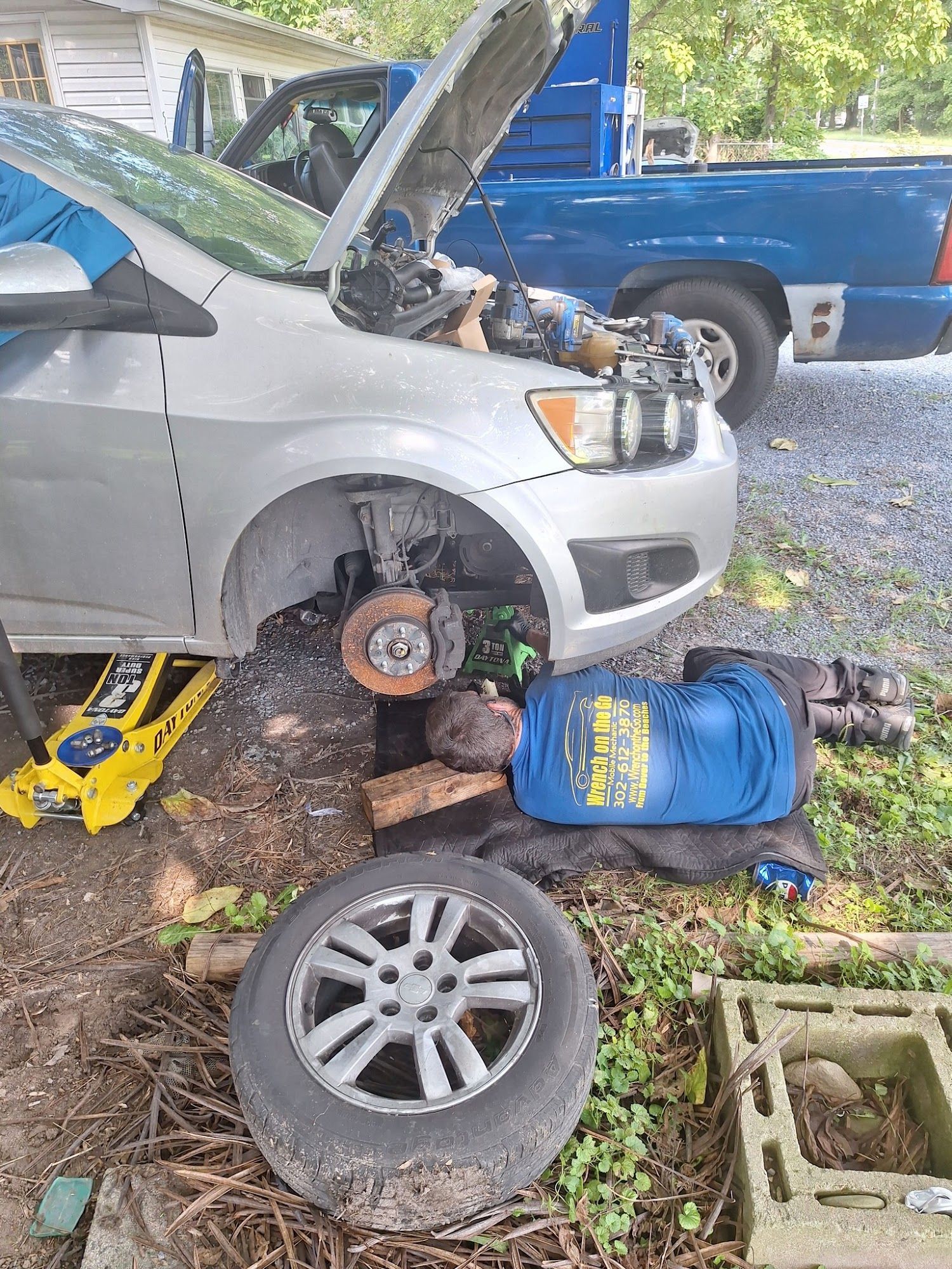 Silver car with open hood, tire off, jack in use, tools, and blue truck in a yard.