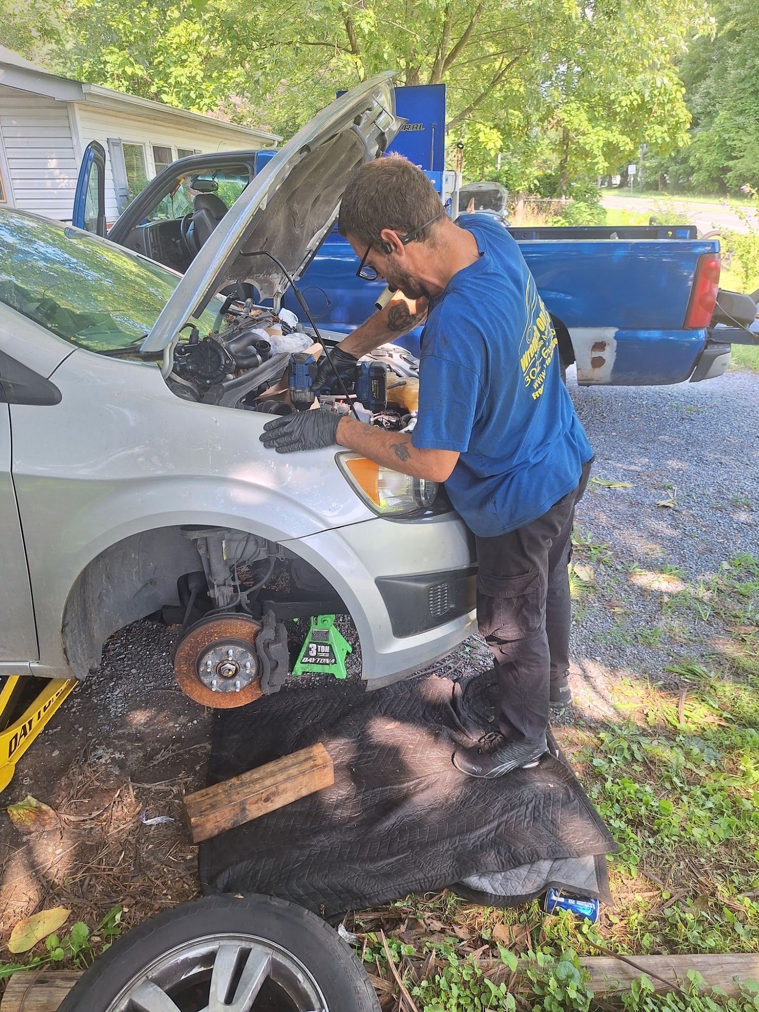 Man working on a car with the hood open, outdoors. Car is raised on a jack. A blue pickup truck is in the background.