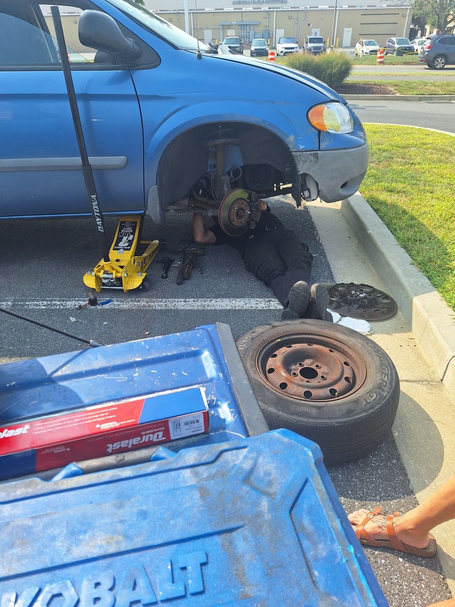 Person working on a blue minivan, tire removed. Tools and parts are visible in a parking lot.