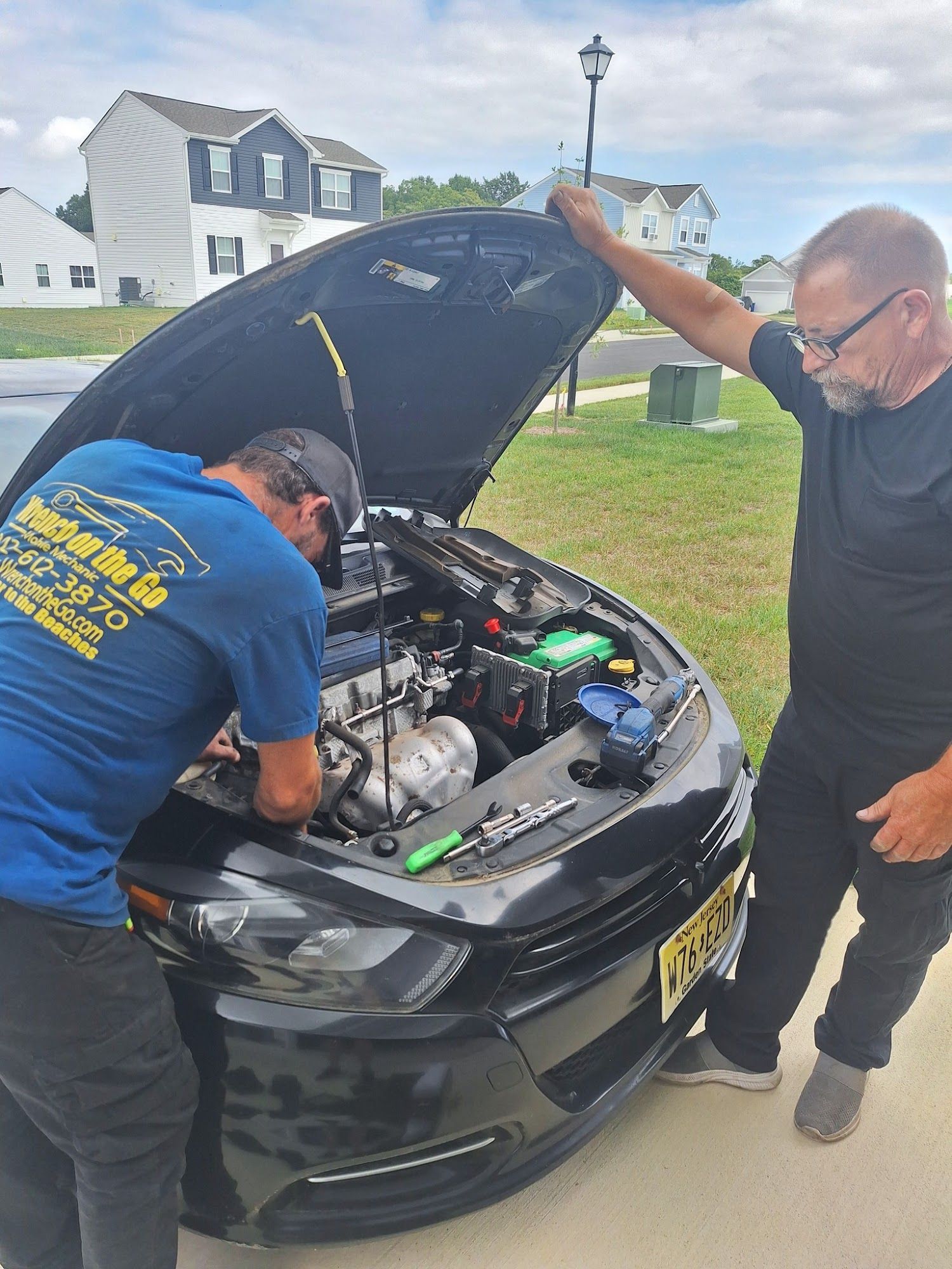 Two men working on a black car with the hood open outside. One man is working, the other is leaning on the car.