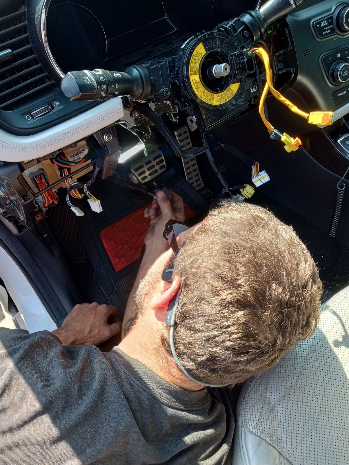 Man working on car's dashboard. Steering wheel and wires exposed. Red mat visible.