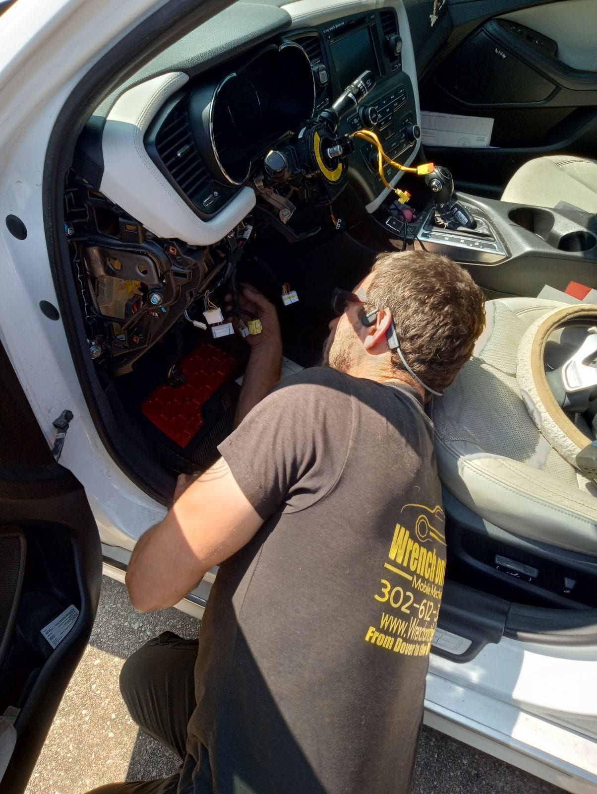 Mechanic working on car's dashboard. Interior of white car with wires exposed. Man wearing t-shirt kneels inside.