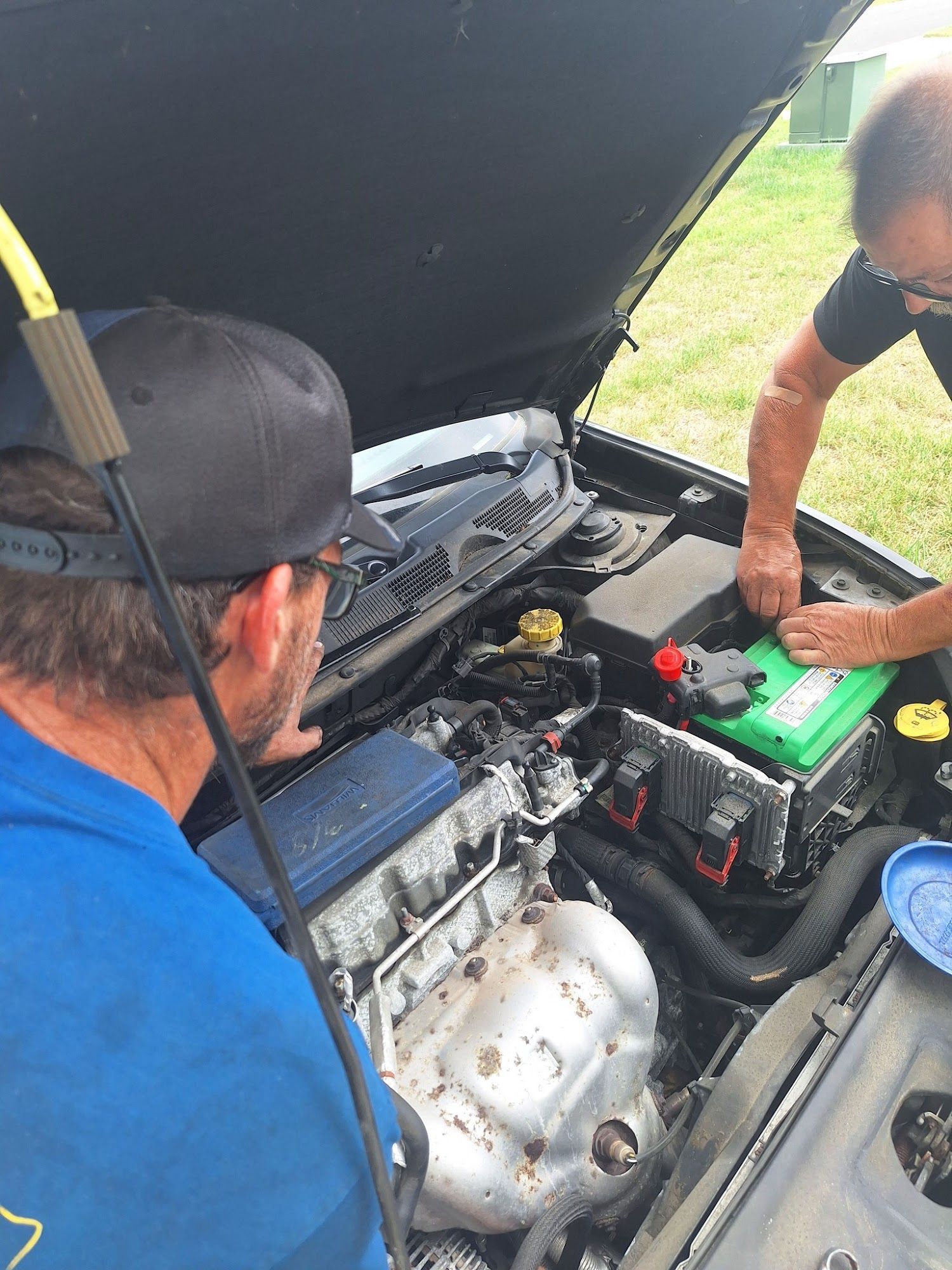 Two men working on a car engine under the open hood; one holds a tool, the other adjusts something near the green battery.