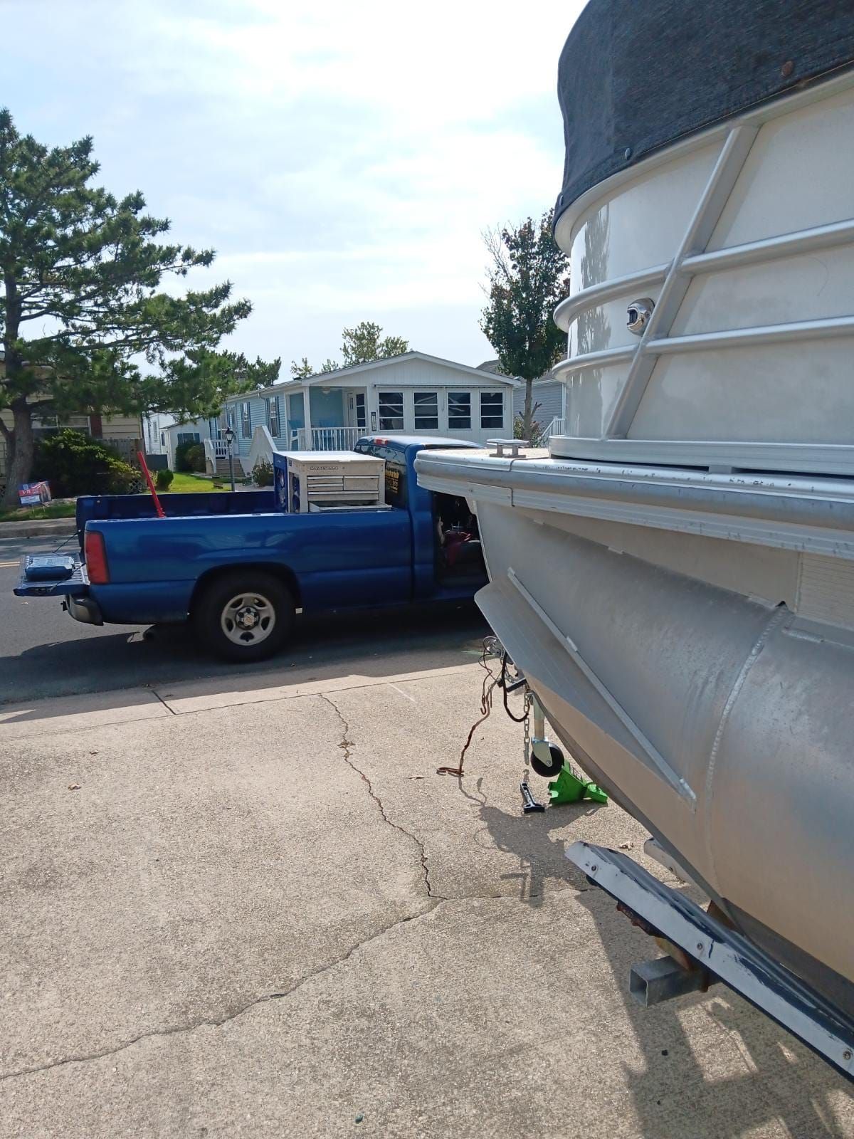 Blue pickup truck next to a pontoon boat, parked on concrete in front of houses.