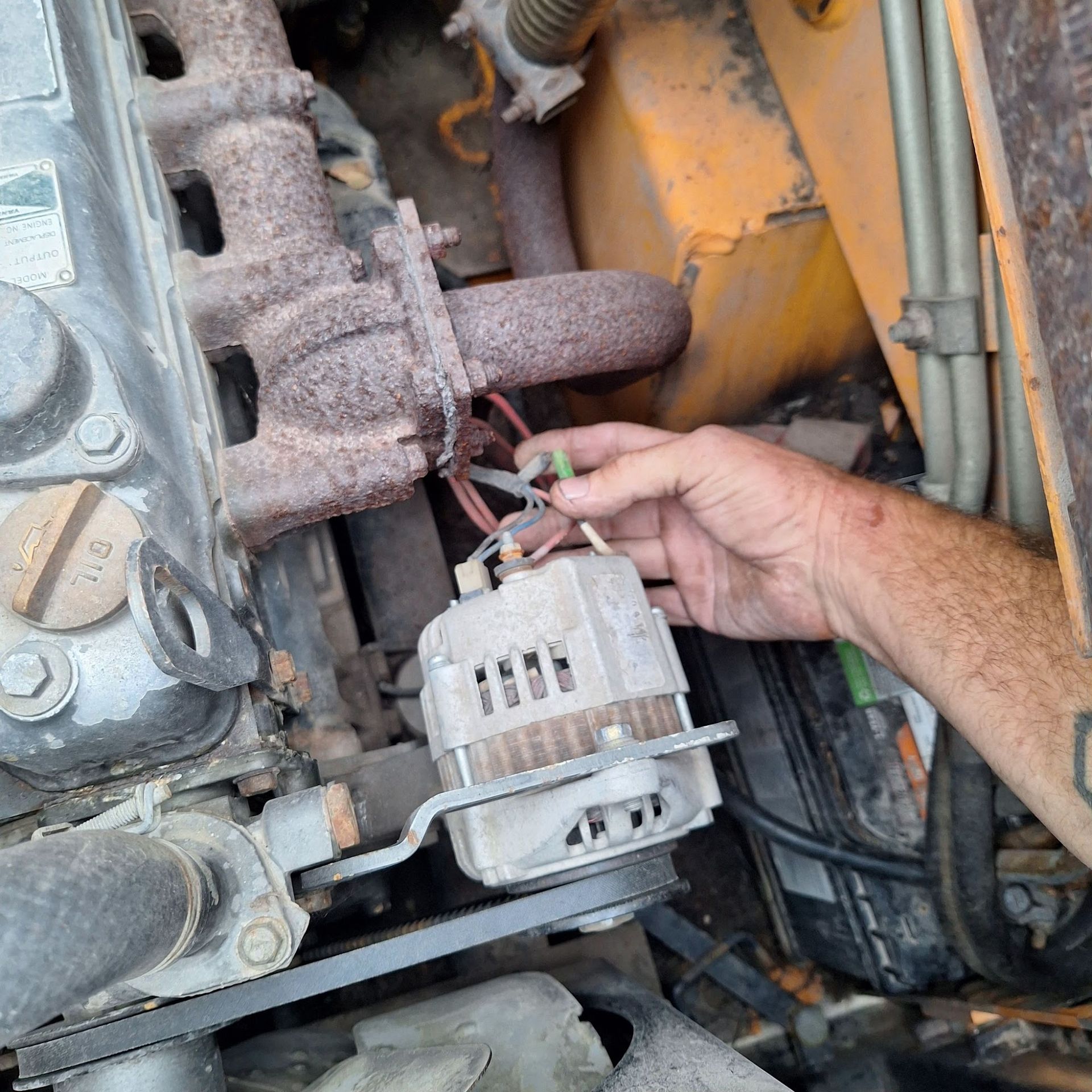 Hand holding an alternator near an engine. Wires and a rusty exhaust manifold are visible in an engine compartment.