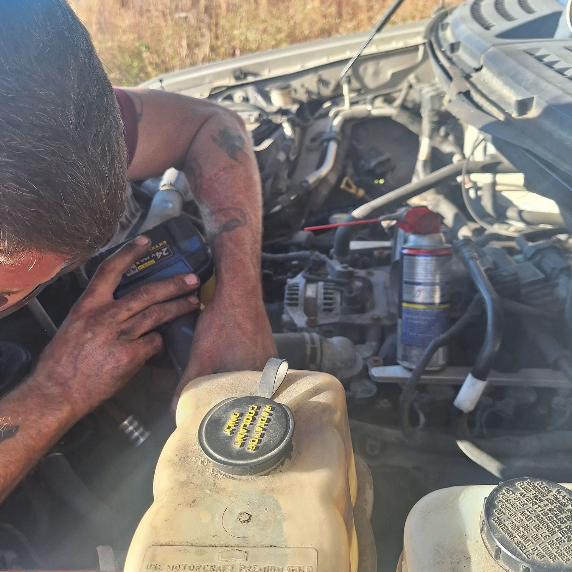 Person working on a vehicle's engine bay. Dirty hands, tools visible. Yellow coolant reservoir cap in foreground.