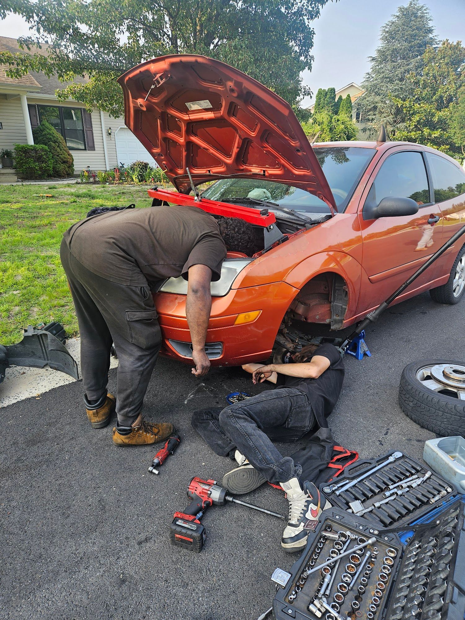 Two people working on an orange car with the hood open in a driveway. Tools are scattered nearby.