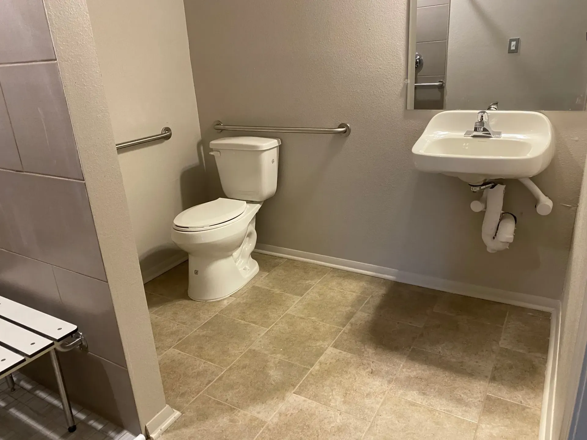 Accessible public restroom with toilet, sink, grab bars, and a bench. Light-colored walls and flooring.