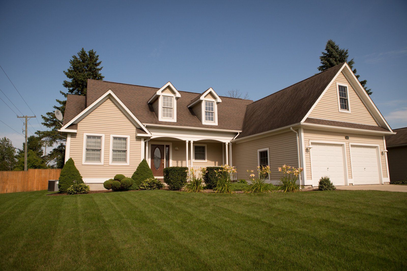 House with beige siding, brown roof, three dormer windows, and a two-car garage under a blue sky.