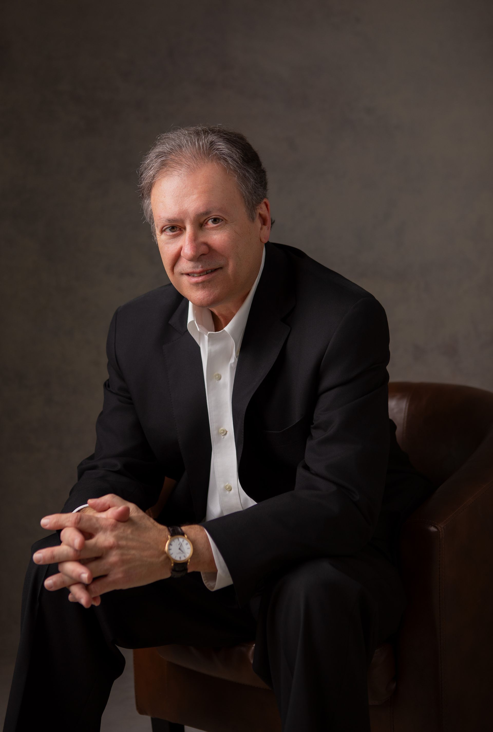 Man in a black suit and white shirt sitting in a chair, hands clasped.