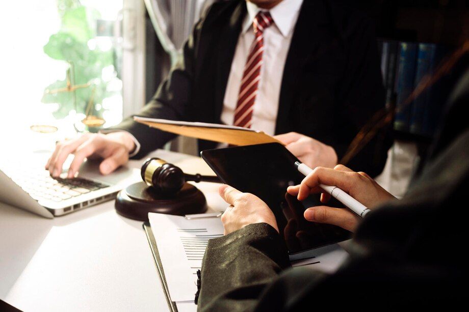 Two people in suits at a desk, reviewing documents and using a laptop; gavel nearby.