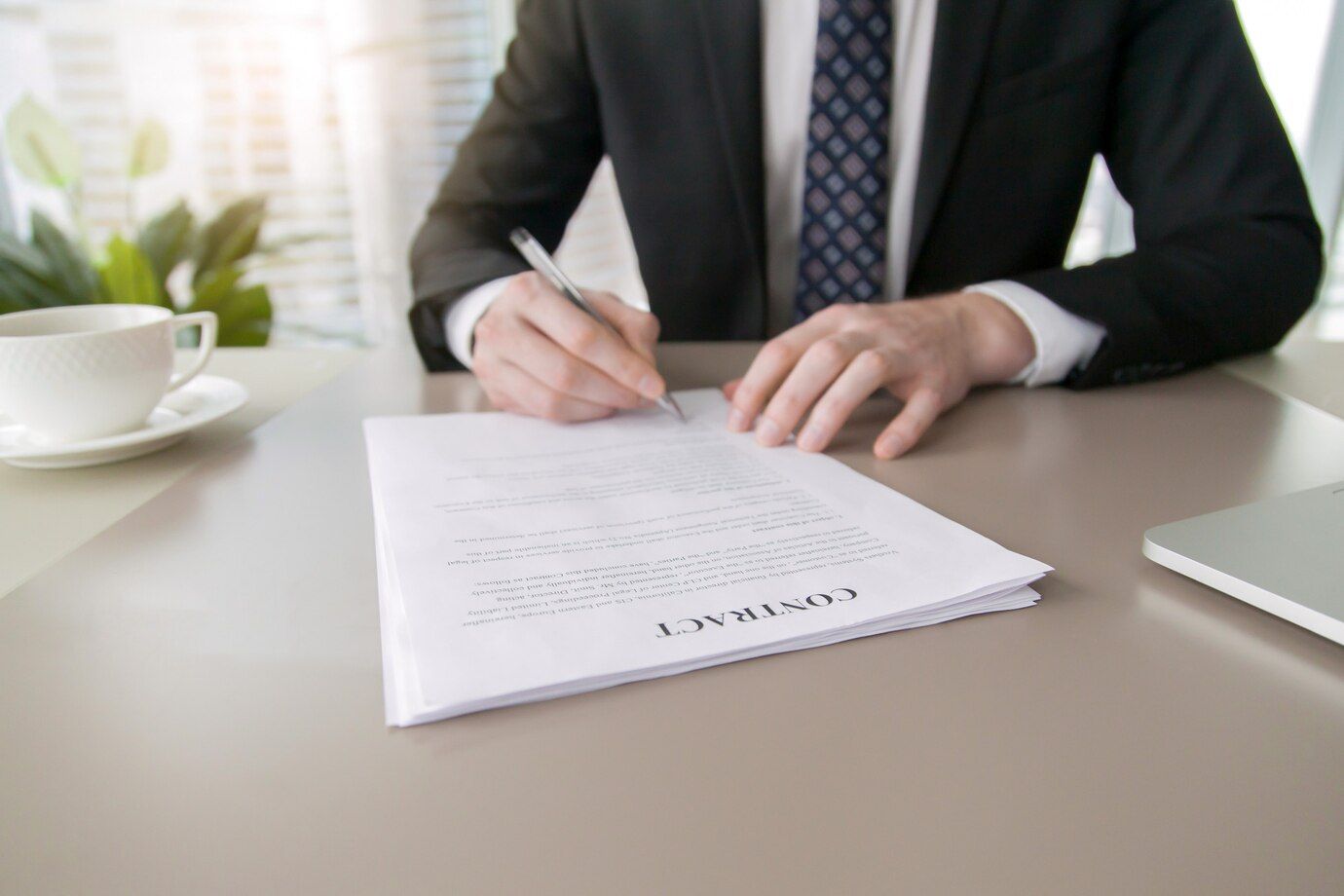 Man in suit signing a contract at a desk with a coffee cup and a laptop.