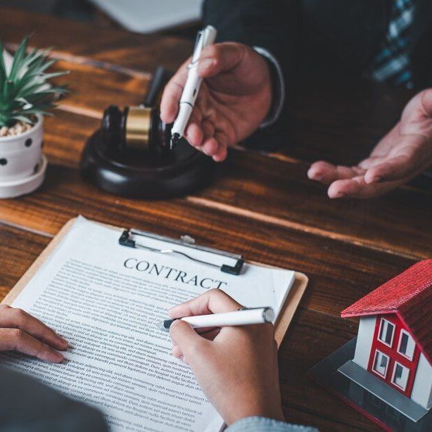 Person signing a contract, with a lawyer, gavel, and toy house on a wooden table.