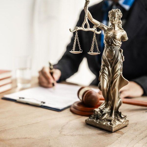 Statue of Lady Justice on desk with person writing. Gavel and legal documents are also present.