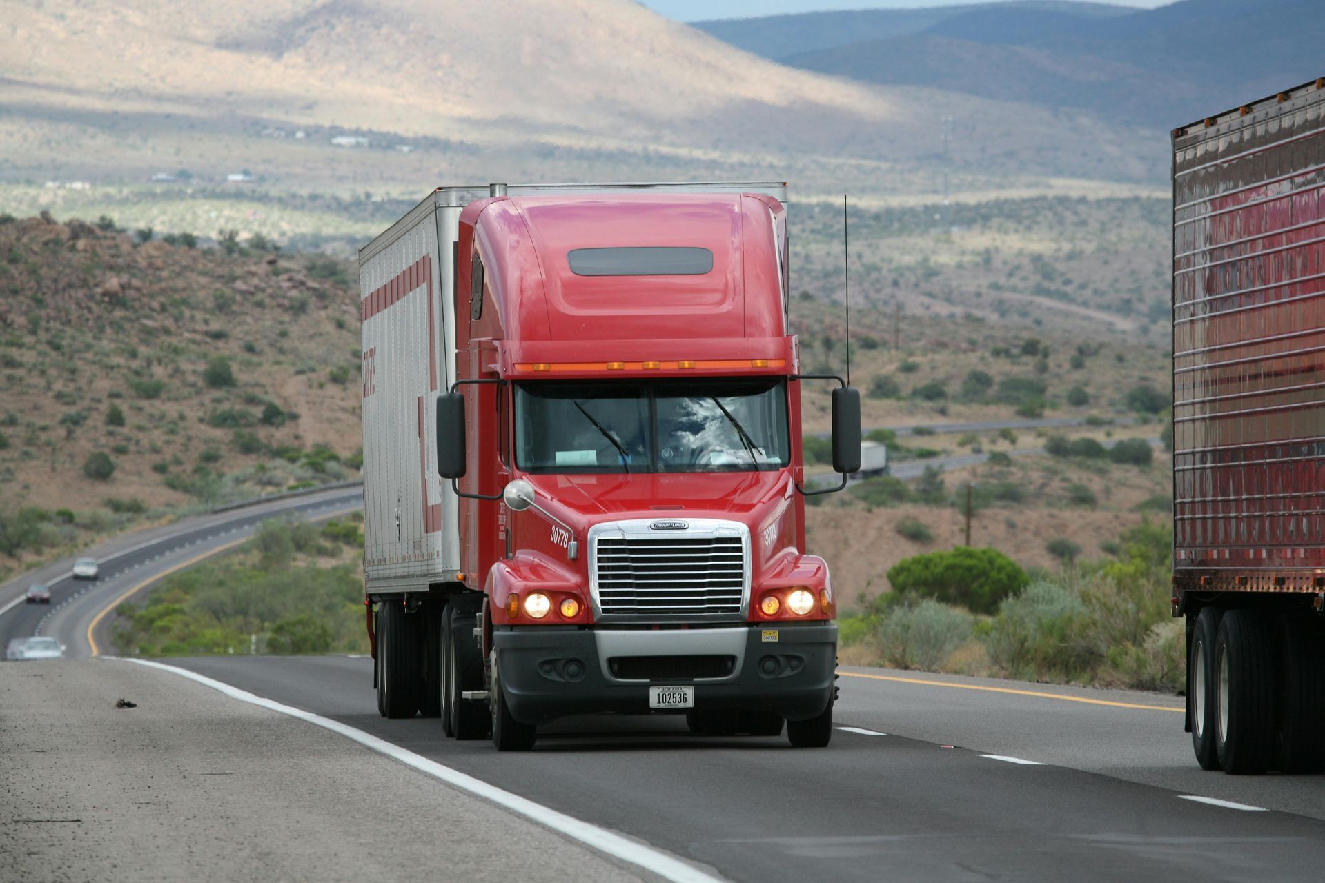 Red semi-truck driving on a highway, another truck to the right. Mountainous landscape in the background.