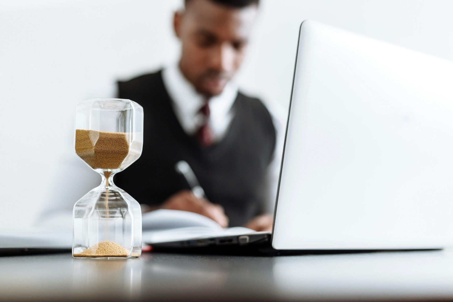 Hourglass on desk, blurred person working on laptop and writing.