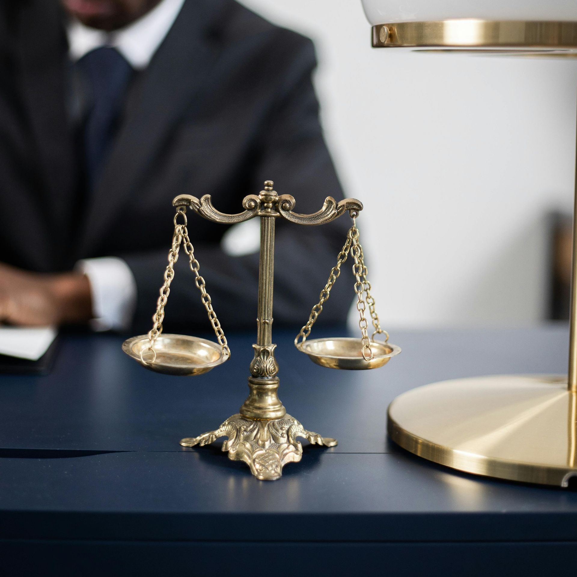 Scales of justice on a desk; a person in a suit is blurred in the background.