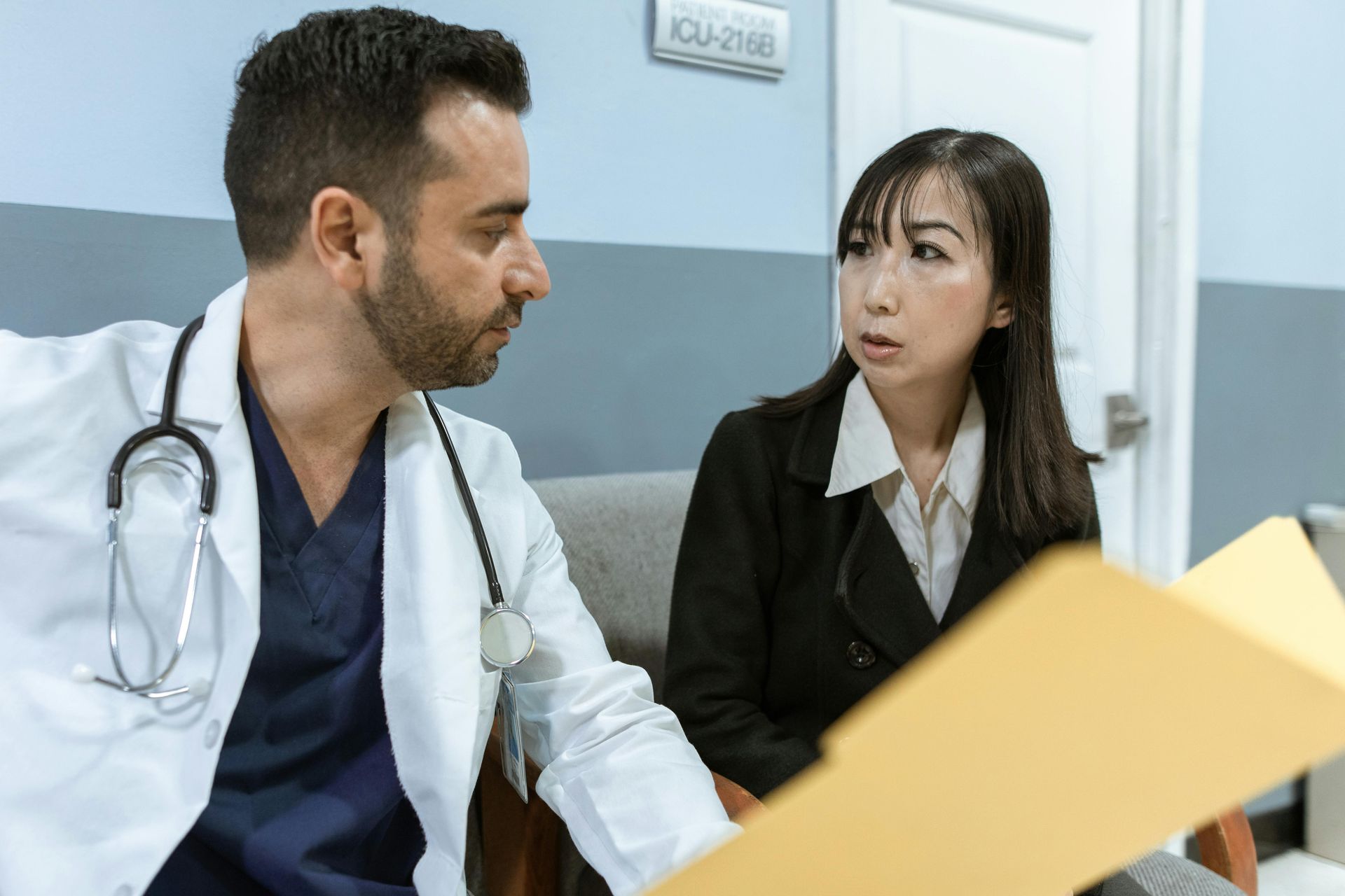 Doctor in a white coat with a stethoscope reviews a file with a concerned patient in a blazer in a waiting room.