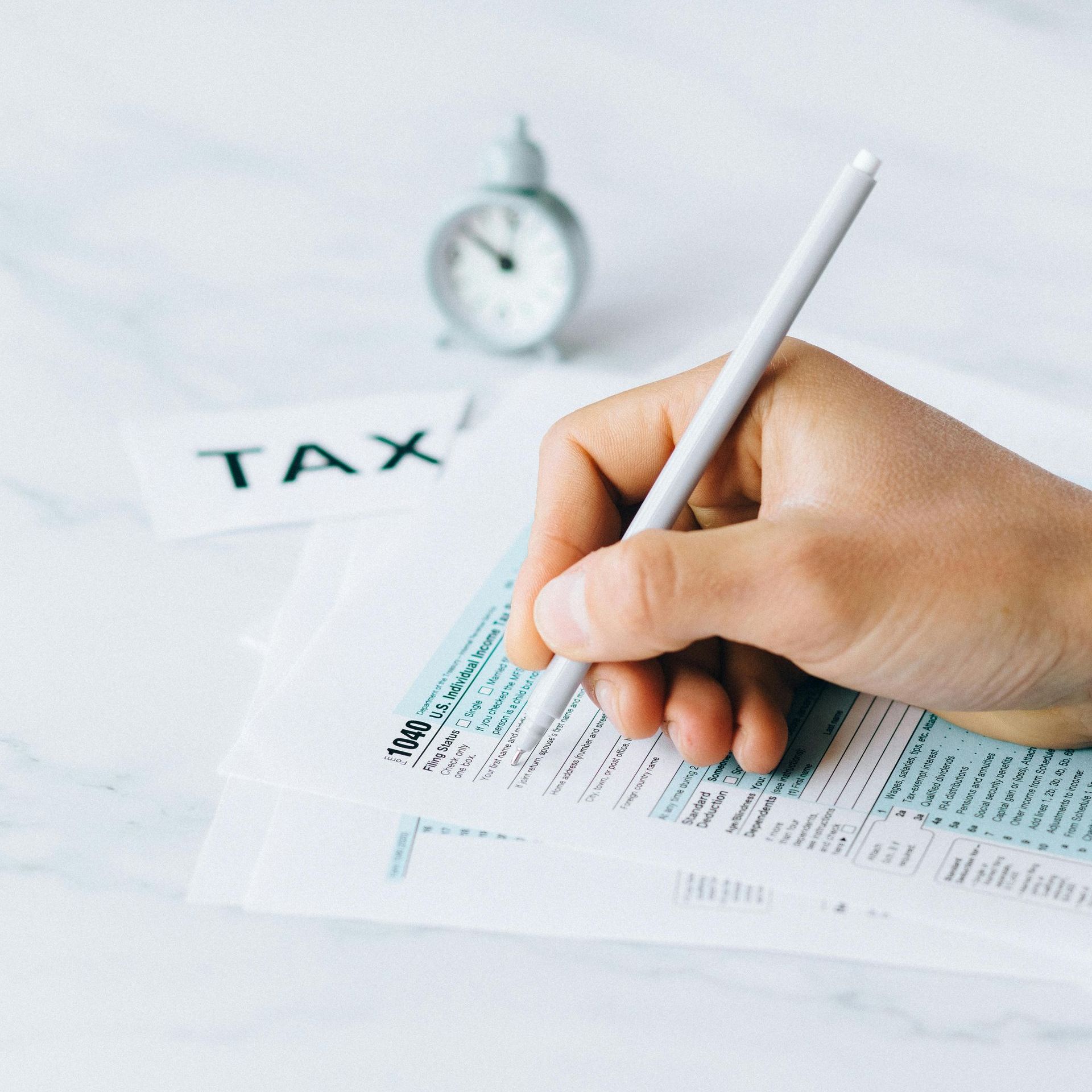 Hand filling out tax forms with a pen; small clock and