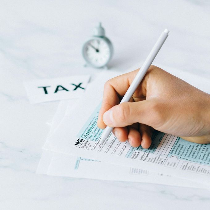 Hand filling out tax forms with a pen; small clock and 