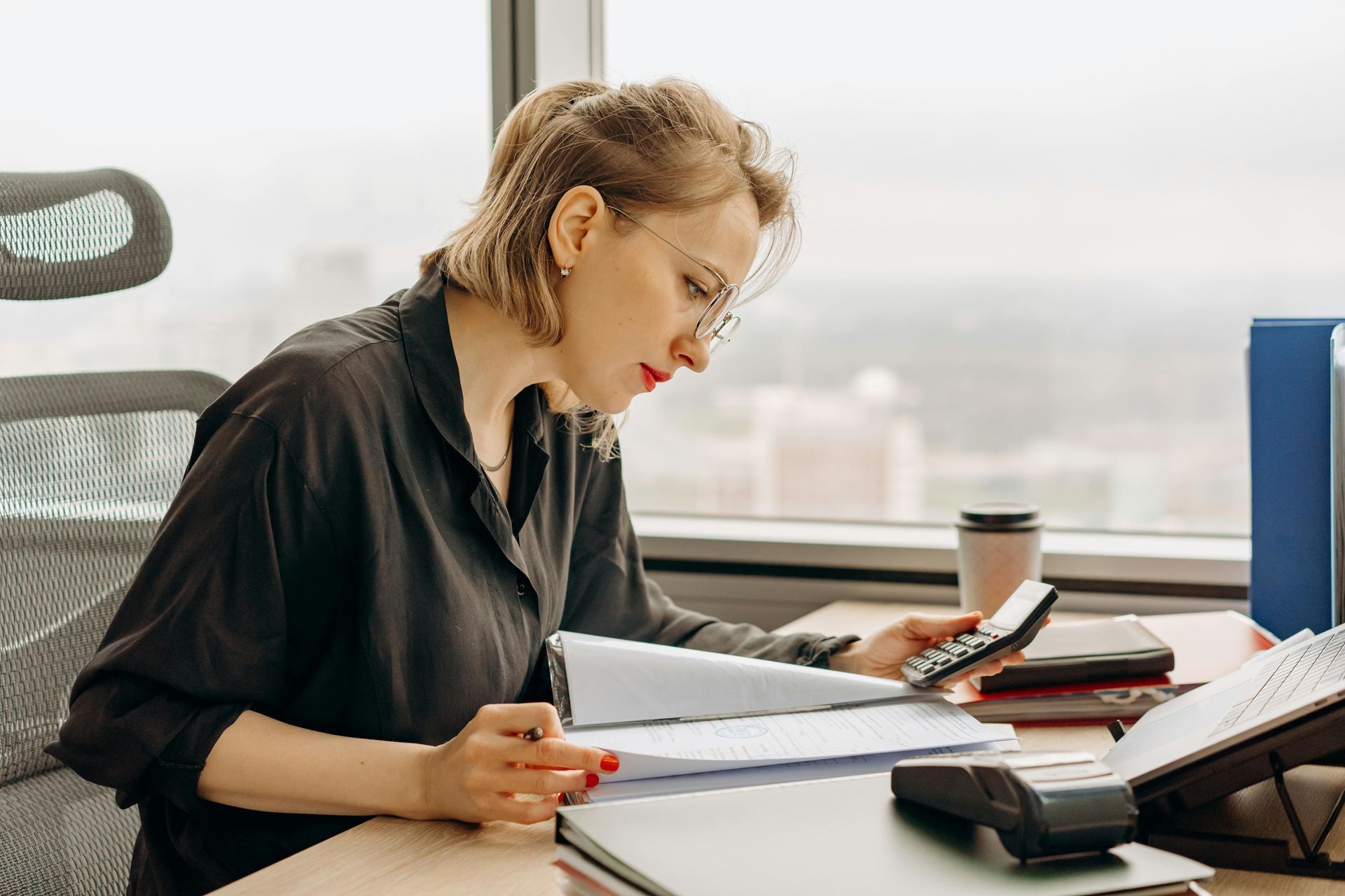 Woman using calculator, reviewing documents at a desk in an office setting.