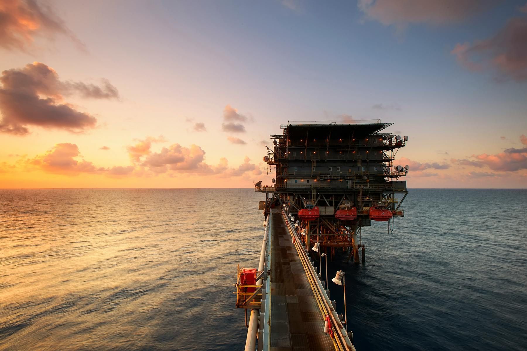 Oil platform at sunset, connected to a pier extending into the sea; orange and purple sky.